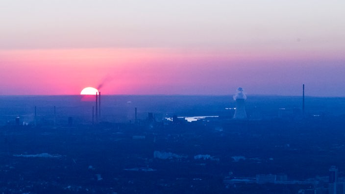 Die Sonne geht rot glühend am Horizont unter, im Vordergrund Industrieanlagen.