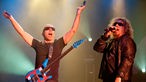 Joe Satriani hat seine Gitarre um den Hals hängen und wirft die Hände in die Luftrechts neben ihm Sänger Sammy Hagar am Mikrofon