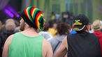 Rückansicht von zwei Festivalbesuchern, der eine mit Mütze in Jamaika-Farben, der andere mit einem Jamaika-Cap.