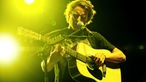 Ben Howard steht mit seiner Akkustikgitarre am Mikrofon und wird von gelbem Scheinwerferlicht umgeben