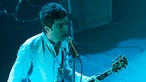 Ein in blaues Licht getauchter Noel Gallagher steht auf der Bühne.