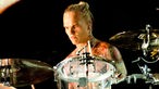 Schlagzeuger mit langen zu einem Zopfg gebundenen Haaren, nacktem Oberkörper und vielen Tatoos auf dem Körper spielt mit einem ernsten Gesichtsausdruck ein Schlagzeug