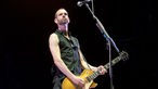 Gitarrist mit schwarzem Shirt, kurzem Haar und 3-Tage-Bart spielt eine gelbliche Gitarre; im Vordergrund steht ein Mikrofon