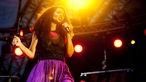 Joy Denalane steht in einem lila Rock auf der Bühne. Sie hält in der rechten Hand den Mikrofonständer, während sie in der linken Hand das Mikrofon hält. 