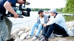 Patrice sitzt mit Interviewer Manuel Unger am See und wird dabei von einem Kameramann gefilmt.