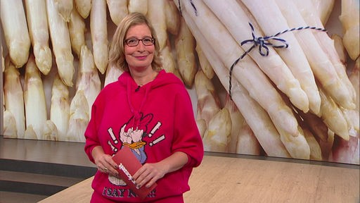 Das Bild zeigt Yvonne Willicks im Fernsehstudio. Im Hintergrund ist ein Bild von verschiednene Spargelbündeln zu sehen.
