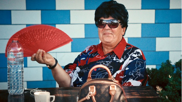 Eine Frau mit Sonnenbrille und Fächer sitzt an einem Tisch