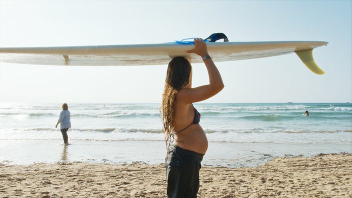 Eine schwangere Frau am Strand trägt ein Surfbrett