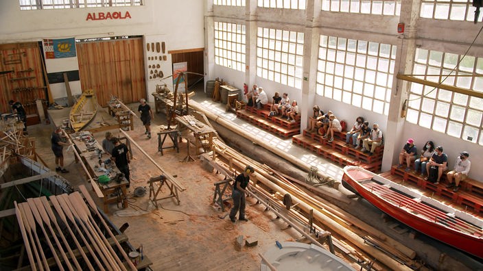 Mehrere Handwerker in großer Werkstatt bei Holzarbeiten werden von einem Dutzend Zuschauern beobachtet