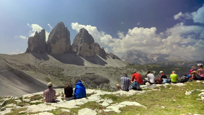 Mehrere Wanderer sitzen an einer Bergkante und schauen auf gegenüberliegendes Bergmassiv mit drei Bergspitzen
