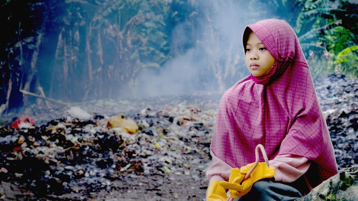 Ein Mädchen mit gelben Gummihandschuhen sitzt vor einer Landschaft voll von Plastikmüll